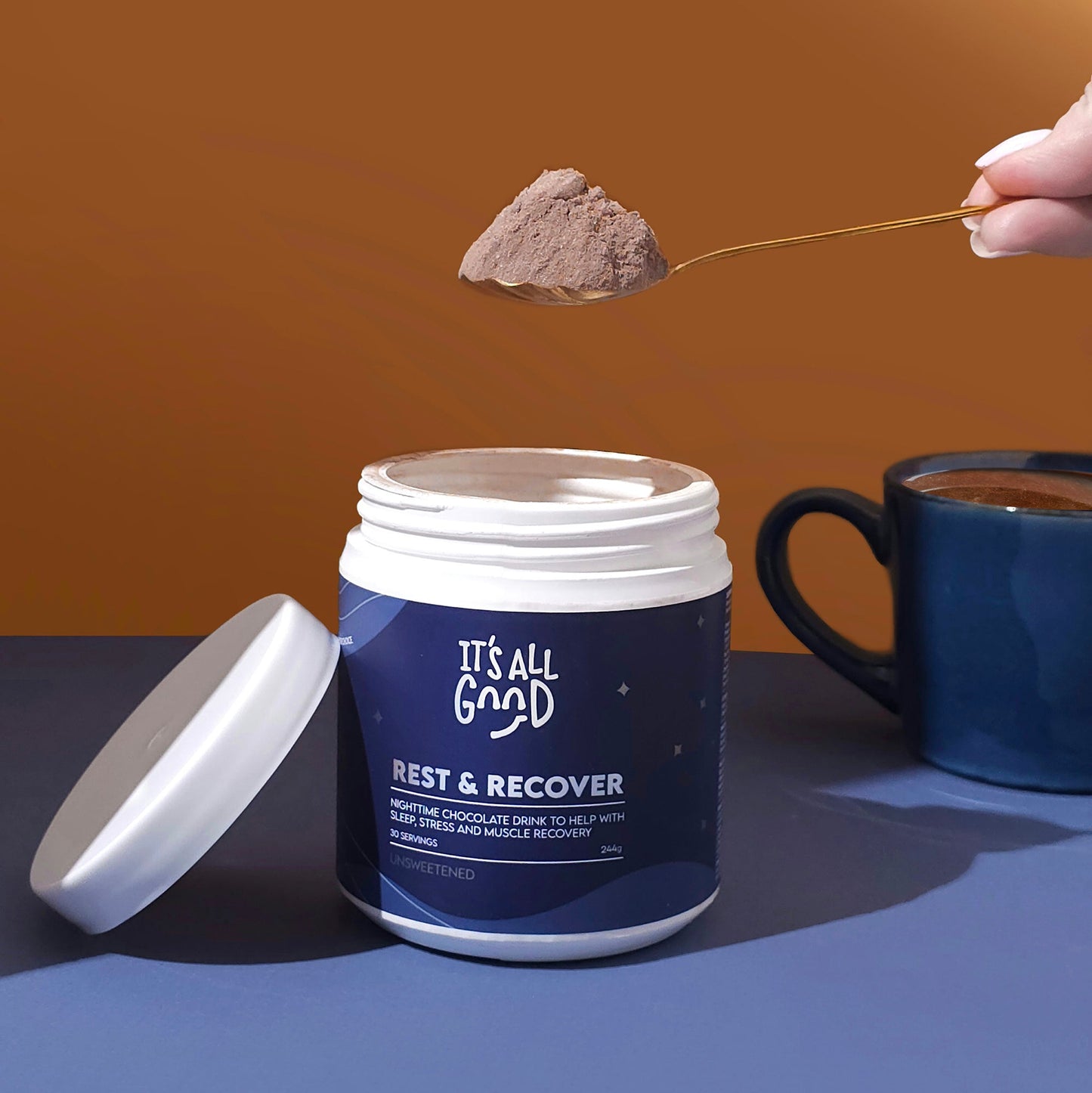 Container of 'It's All Good Rest & Recover' powder with a spoonful being lifted, on a blue surface with a brown background.