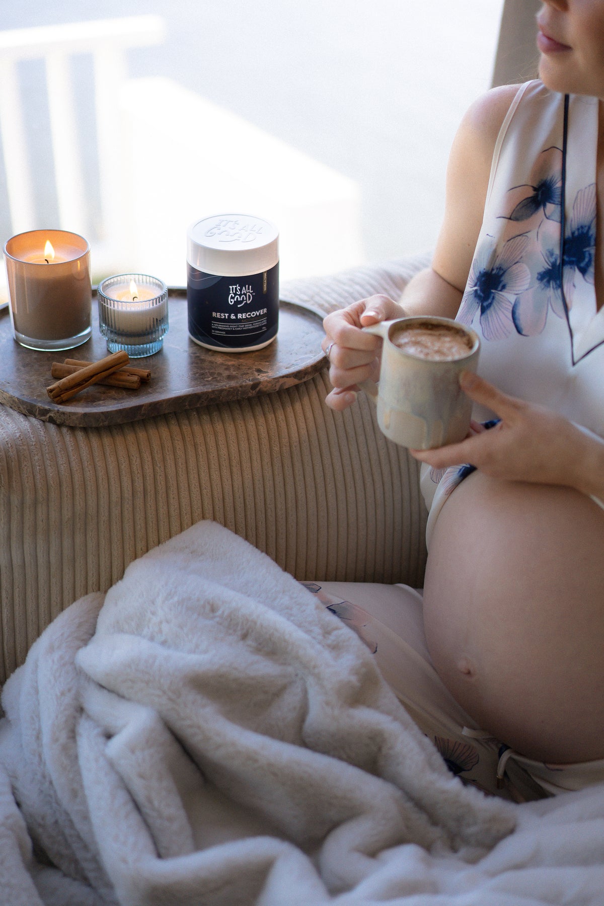 A cozy lifestyle scene featuring a pregnant woman holding a mug of hot cocoa while sitting on a ribbed beige couch. A wooden tray on the couch holds a lit candle, a small glass candle, cinnamon sticks, and a dark jar labelled "Rest & Recover"