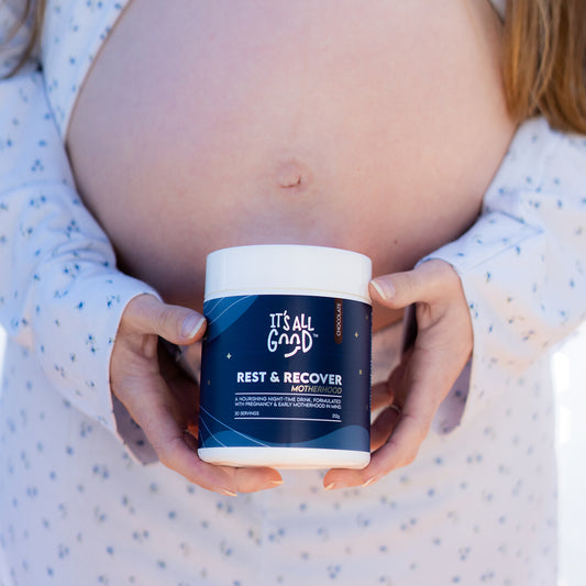 A pregnant woman holds a white jar of 'It's All Good' Rest & Recover Motherhood against her stomach. She is wearing a white long-sleeve pyjamas with small blue star patterns, and the background is a soft, out-of-focus light gray.