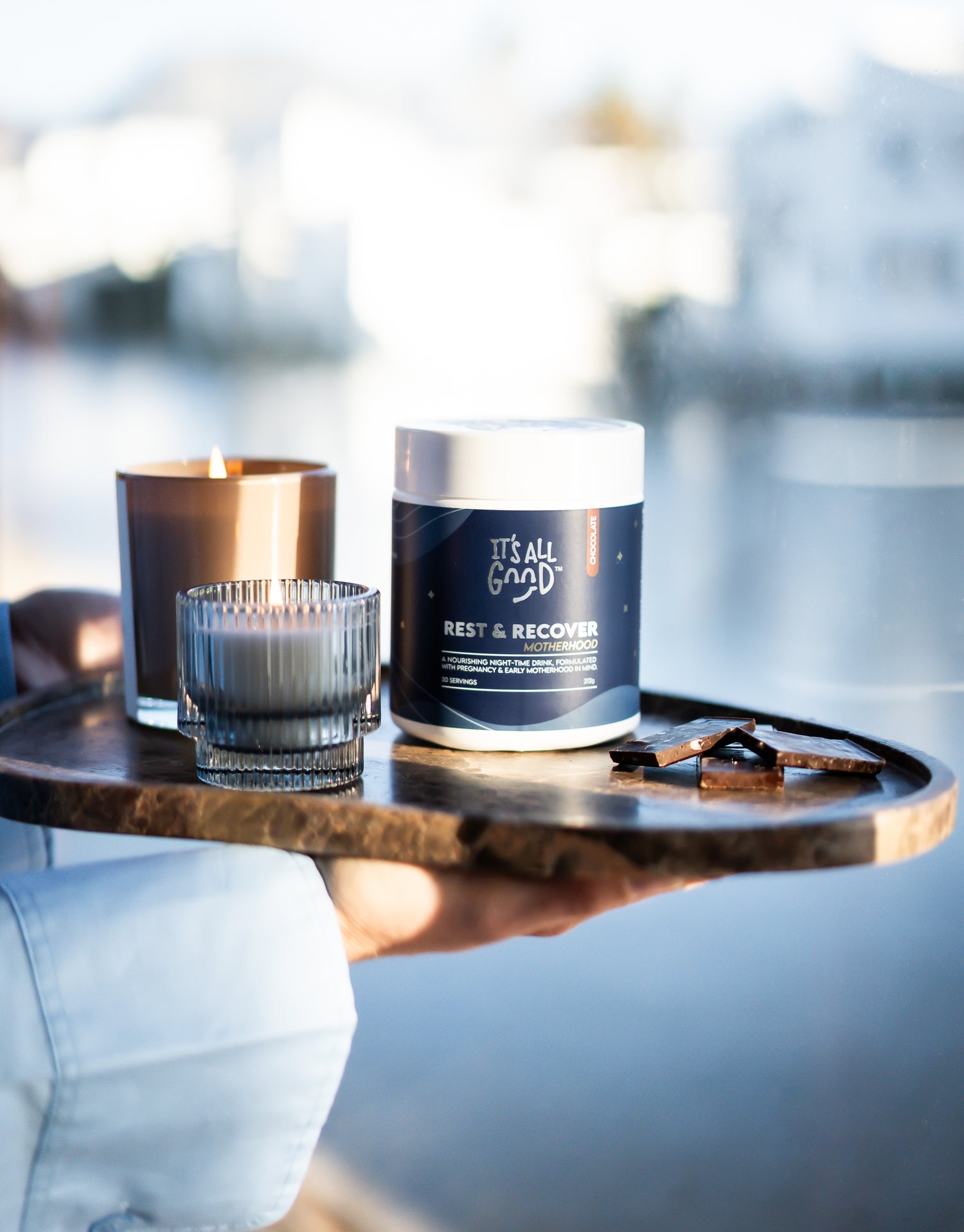 A hand holding a wooden tray with a jar candle labeled 'IT'S ALL GOOD REST & RECOVER' in navy and white packaging, alongside a lit amber glass candle and a small ribbed glass candle holder, against a blurred outdoor waterfront background.