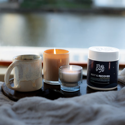 A tub of Rest & Recover Motherhood by It's All Good, styled on a dark tray with a ceramic mug, a lit candle, and a small glass candle holder in front of a window overlooking water.