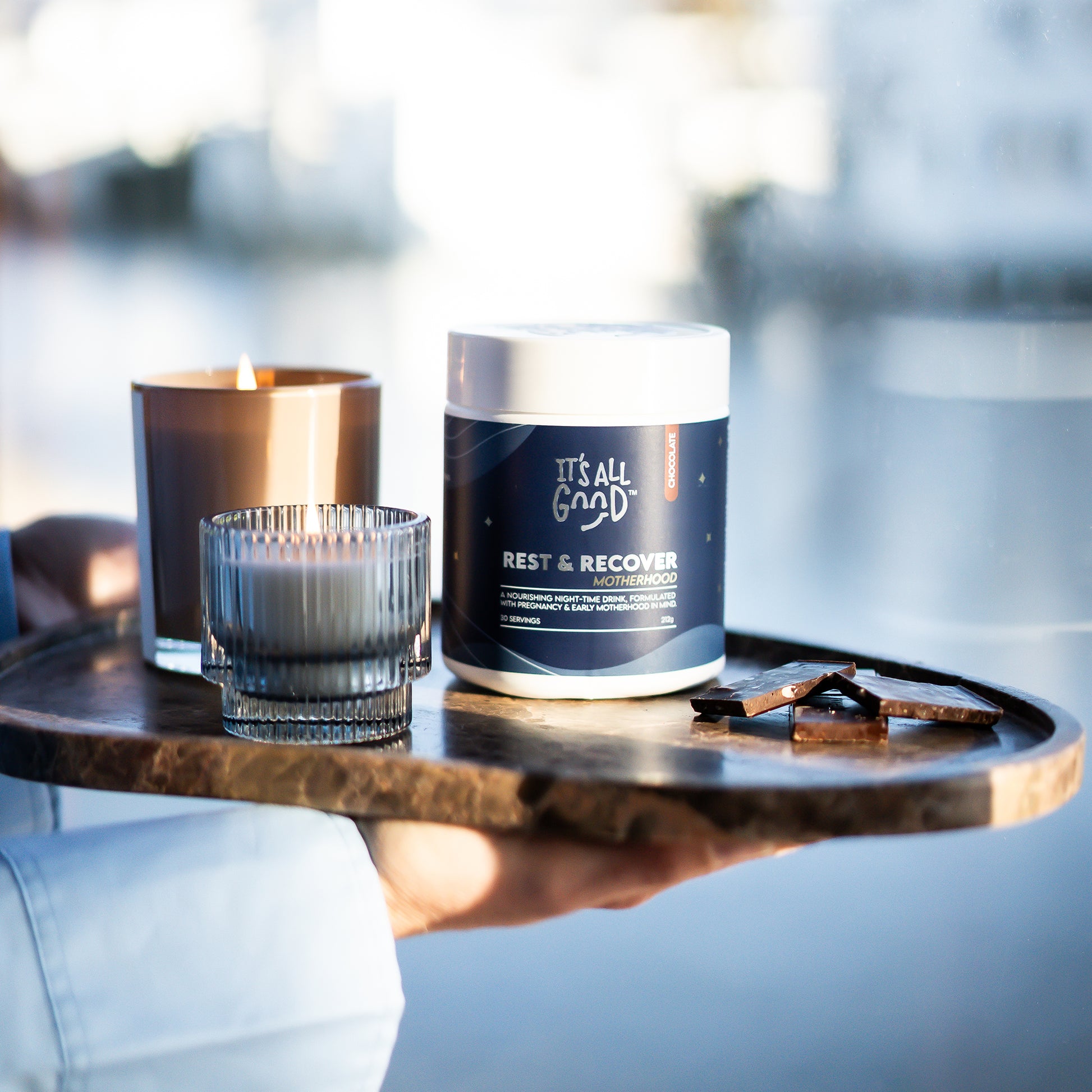 A hand holds a round wooden tray in an outdoor setting with a blurred water background. On the tray are a jar labeled 'IT'S ALL GOOD' Rest & Recover Motherhood, a lit amber candle, a small glass candle holder, and a few dark chocolate pieces.