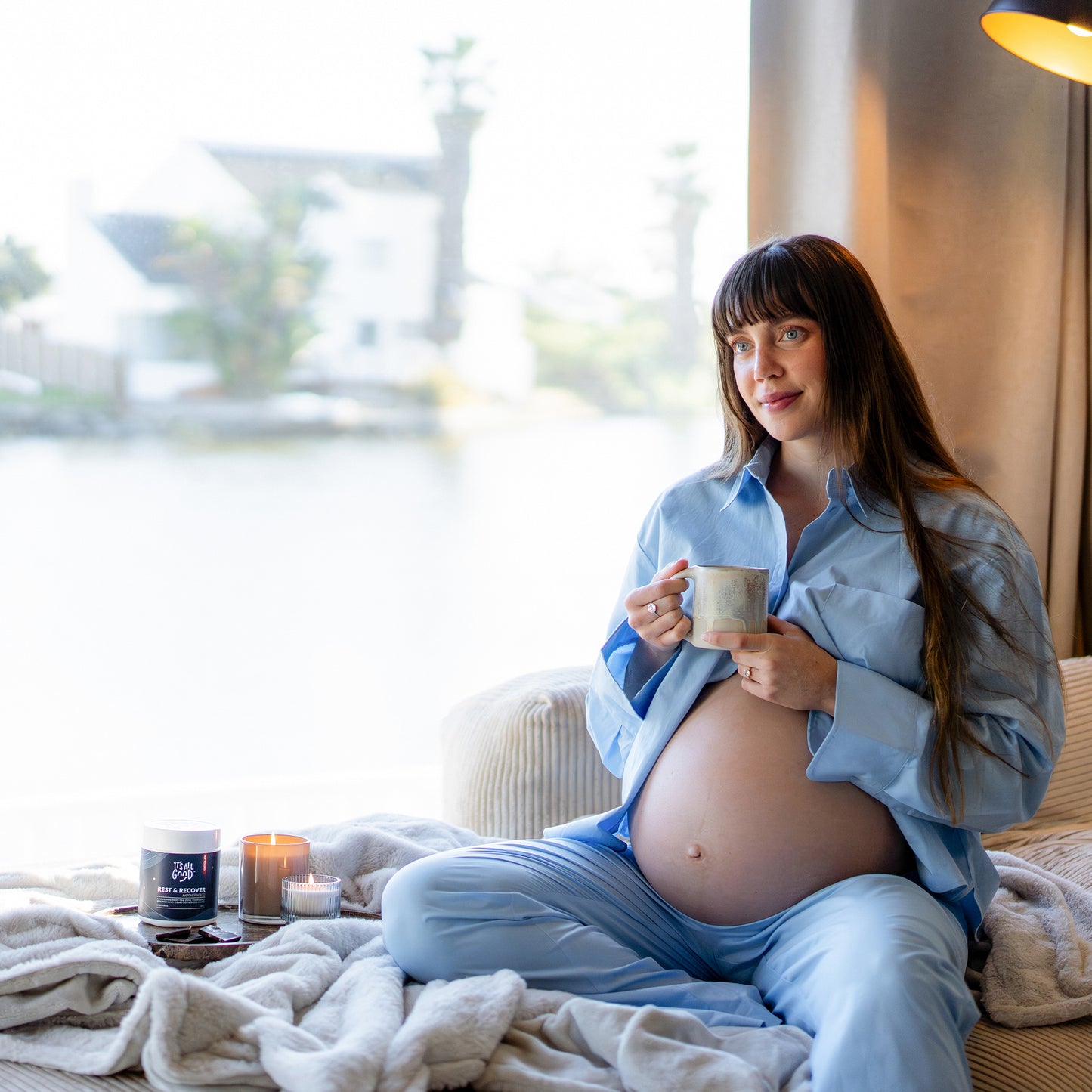A pregnant woman sits on a bed in a bright room, wearing a light blue button-up shirt and matching pants, holding a mug. A small jar and candles sit on the bed beside her.