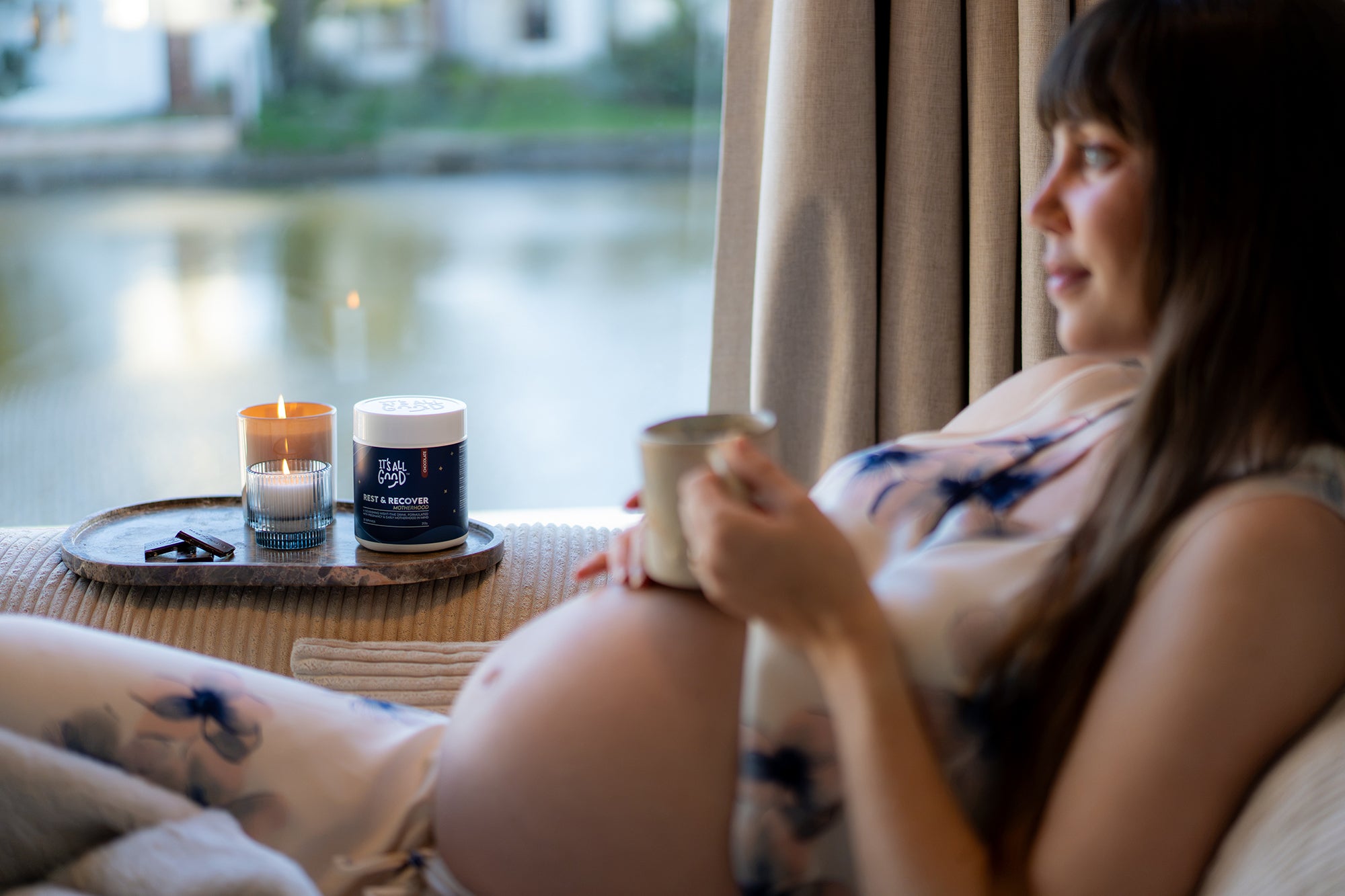 A pregnant woman sits in front of a window overlooking a lake. On the armrest of the couch is a tray with candles and a tub of "Rest & Recover". She has a mug of hot chooclate resting on her belly