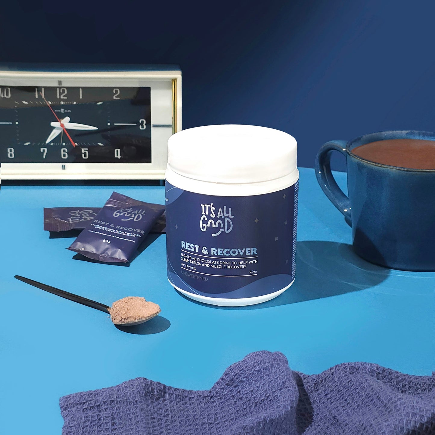 Container labeled 'It's All Good Rest & Recover' on a blue surface with a clock, cup, and packets in the background.