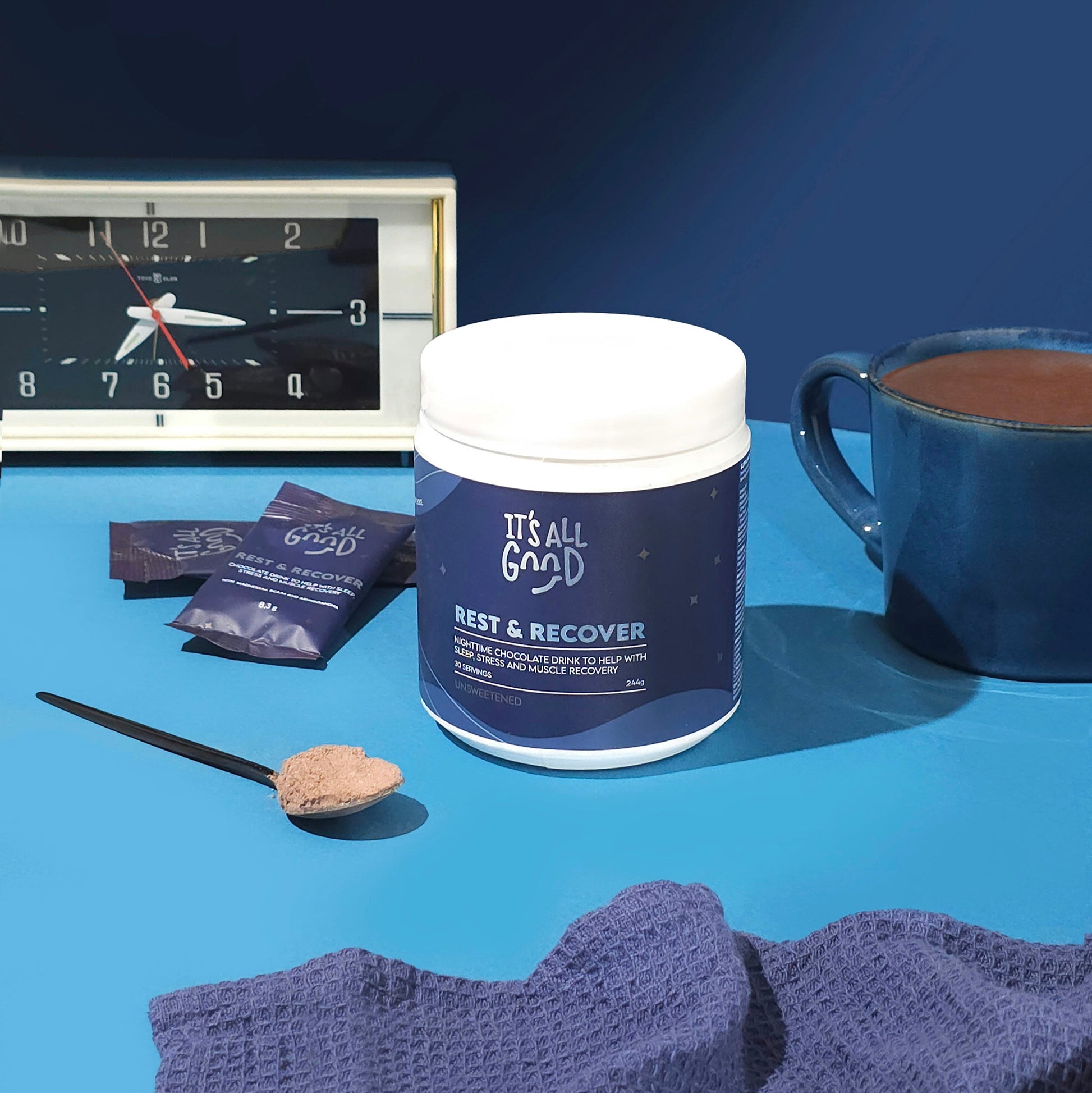 Container labeled 'It's All Good Rest & Recover' on a blue surface with a clock, cup, and packets in the background.