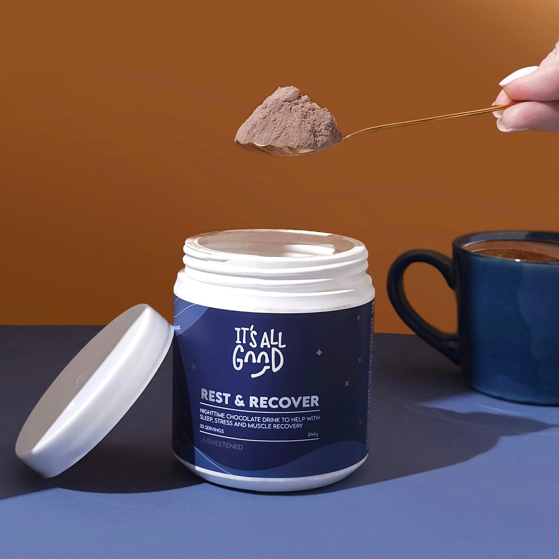 Container of 'It's All Good Rest & Recover' powder with a spoonful being lifted, on a blue surface with a brown background.